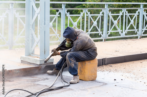 Photos Worker in protective clothes cleans the metal structures sandbla