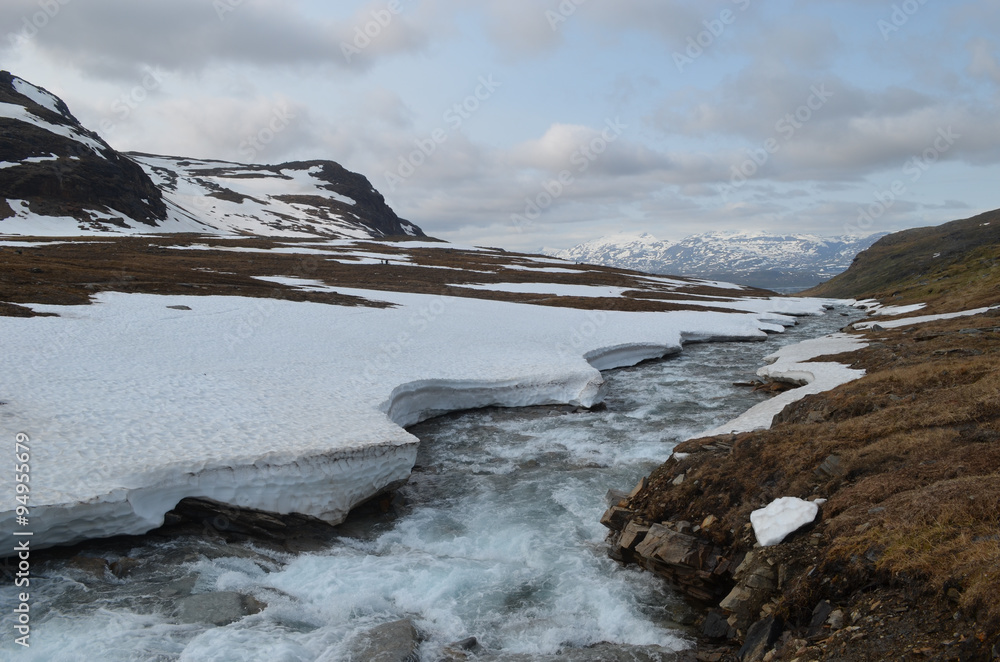 Meltwater river in valley in subarctic mountains with snow on one shore, Lapland
