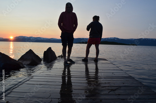 Two silhouets of people standing on pier in Swedish lake with midnight sun photo