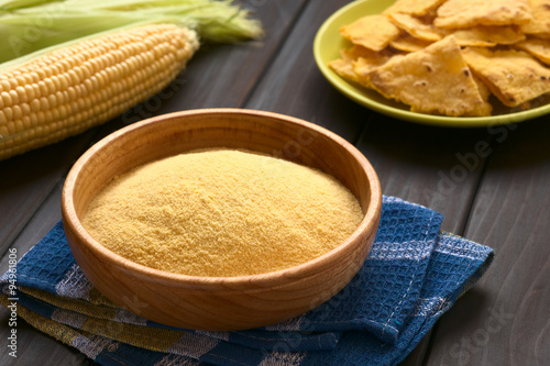 Wooden bowl of cornmeal with homemade tortilla chips and cobs of corn in the back, photographed on dark wood with natural light (Selective Focus, Focus one third into the cornmeal) photo