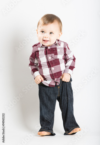 a cute 1 year old baby stands in white studio with jeans and a red white flannel looking camera left in excitement and pulling up his pants