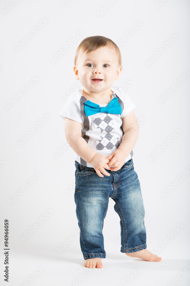 a cute 1 year old stands in a white studio setting. The boy has a happy expression. He is dressed in Tshirt, jeans, suspenders and blue bow tie a cute 1 year old stands in a white studio setting. The boy has a happy expression. He is dressed in Tshirt, jeans, suspenders and blue bow tie