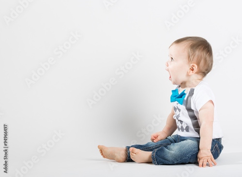a cute 1 year old stands in a white studio setting. The boy is yelling with an open mouth. He is dressed in Tshirt, jeans, suspenders and blue bow tie