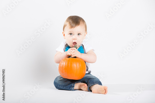 a cute 1 year old sits in a white studio setting with a pumpkin. The boy looks as if he is about to eat the pumpkin stem while looking up. He is dressed in Tshirt, jeans, suspenders and blue bow tie