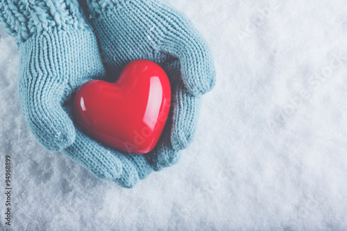 Woman hands in light teal knitted mittens are holding beautiful glossy red heart in snow background. Love, St. Valentine concept photo