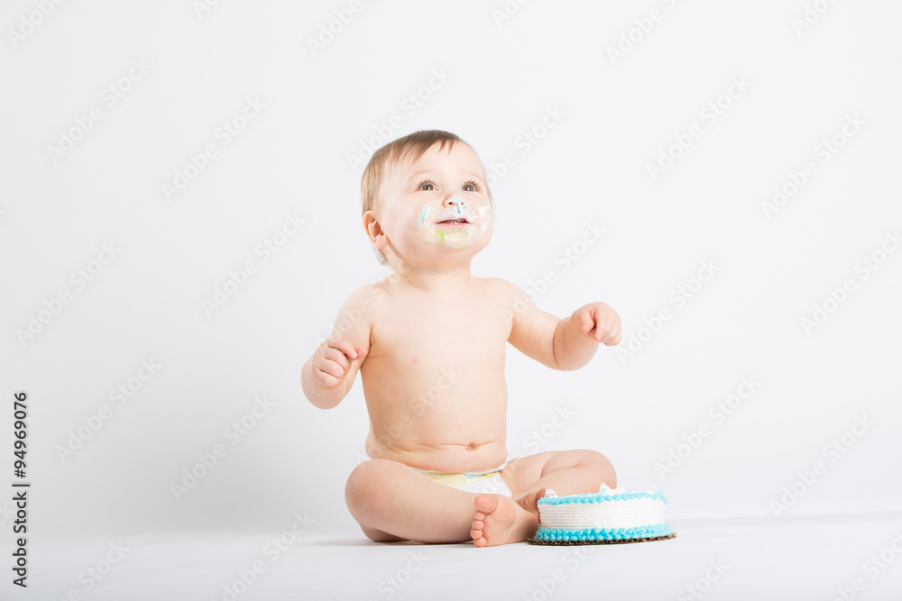 a cute 1 year old sits in a white studio setting. The boy is very excited to start eating his birthday cake. He is only dressed in a white diaper