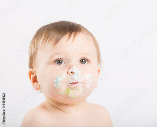a cute 1 year old sits in a white studio setting. Close up of the boys face with cake icing. He is only dressed in a white diaper