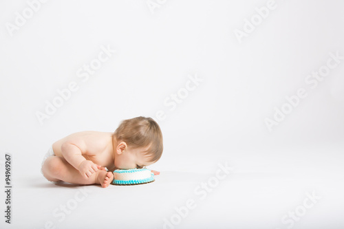 a cute 1 year old sits in a white studio setting. The boy is eating a face full of cake by leaning over into it.copyspace on the right. He is only dressed in a white diaper