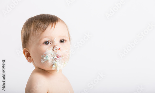 a cute 1 year old sits in a white studio setting. Close up of a baby with a face full of cake and frosting. He is only dressed in a white diaper