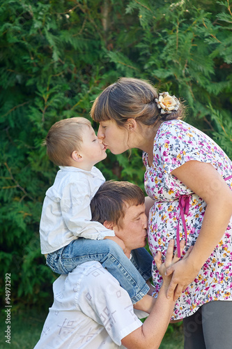 Happy family. Pregnant mother with her husband and son in the park. Mum kisses the son and dad kissing mums belly