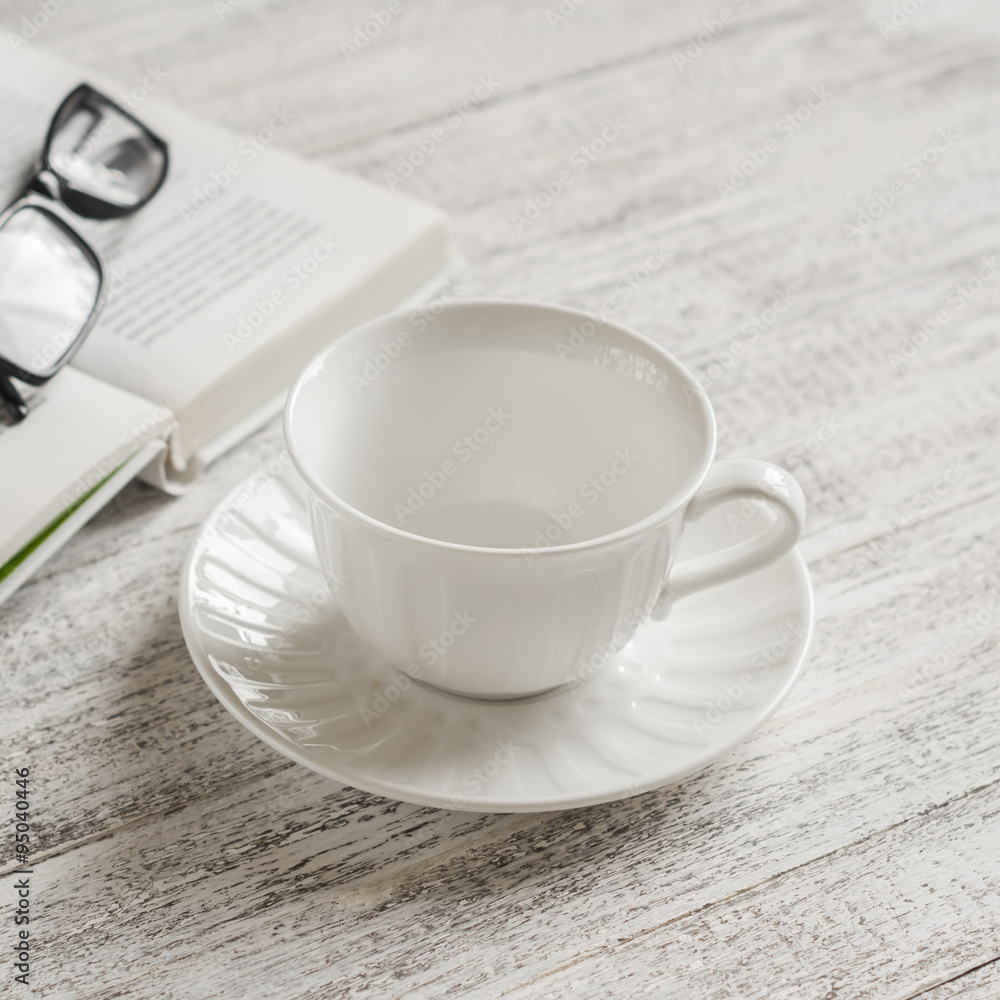 empty white ceramic tea cup, book and glasses on white wooden table