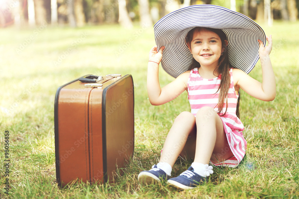 Fototapeta premium Happy little Caucasian girl in striped dress and hat sitting near suitcase, Outdoor portrait