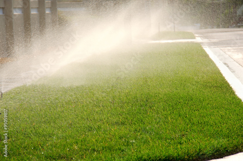 Front yard water sprinkler system watering a field of grass by a residential street.