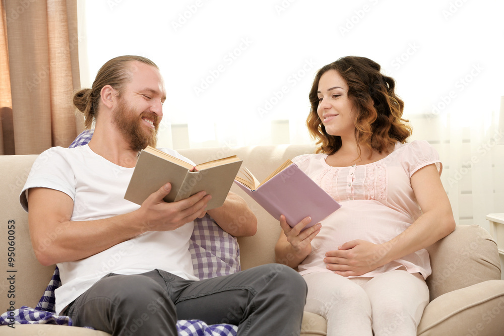 Happy beloved couple reading books in waiting for baby's birth on sofa in the room Happy beloved couple reading books in waiting for baby's birth on sofa in the room