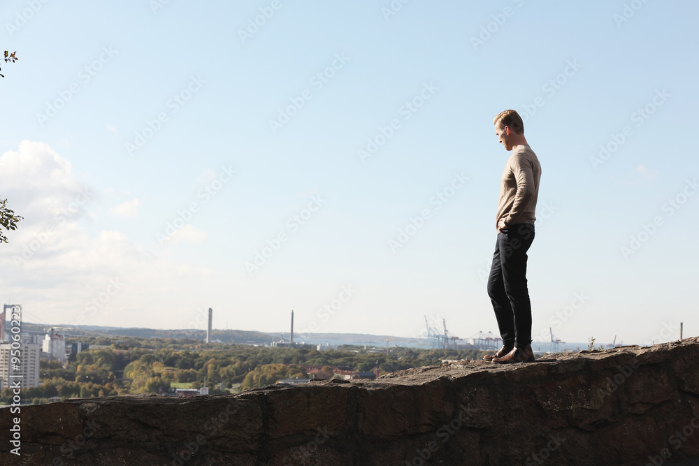 A lonely man wearing a sweater, walking along a stone wall looking at the great view of the city. On a sunny day with blue skys above him.