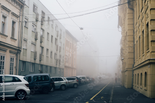 Silent foggy autumn morning street with multi-colored houses bordering the street, cars parked along left side, and electric wires hanging above the street