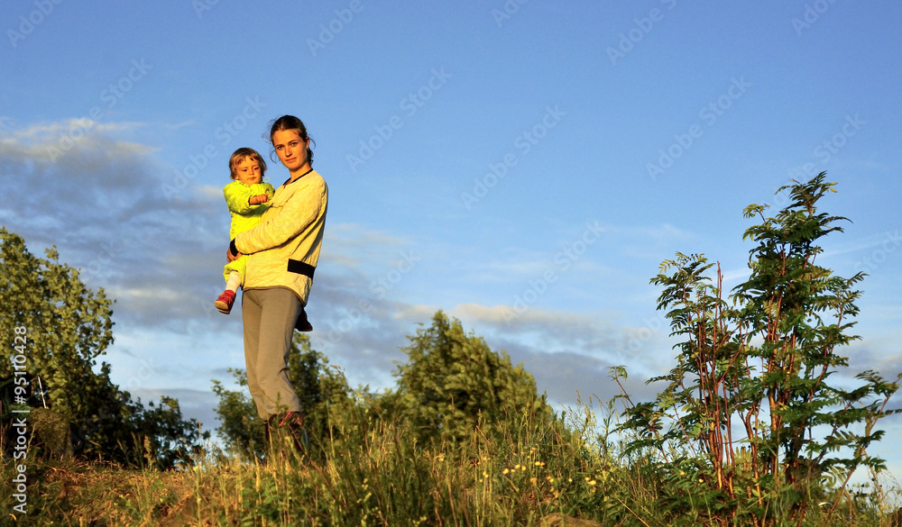 Naklejka premium Young Attractive young mother holding a little girl in her arms at sunset light. Walk in the autumn warm evening outdoors at sunset.