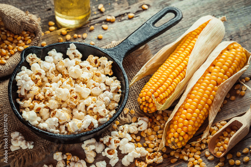Prepared popcorn in frying pan, corn seeds in bowl and corncobs