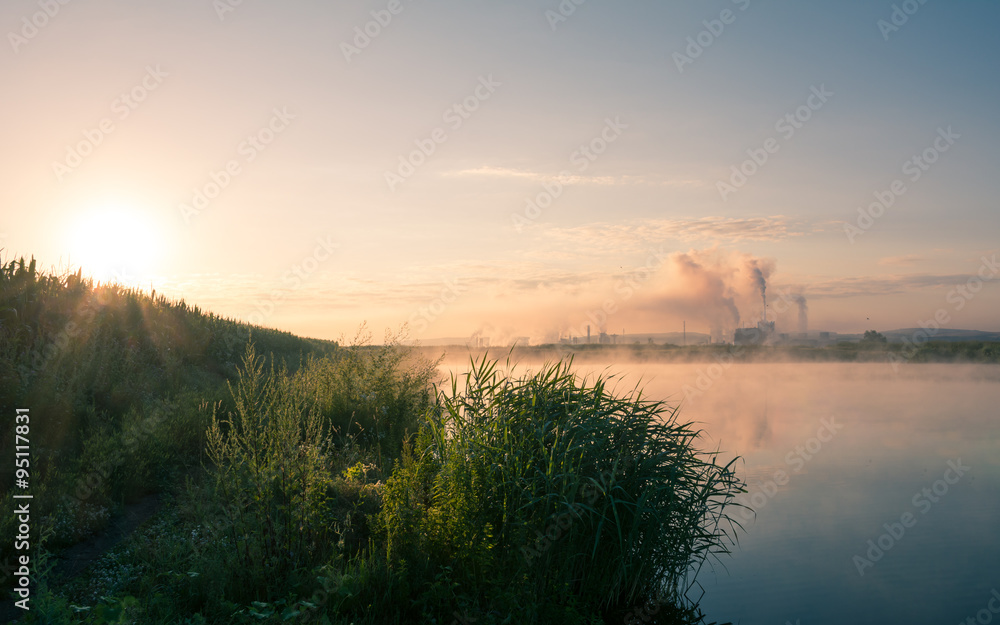 Beautiful early morning mist over a lake and a factory pipe polluting air in the distance. Environmental problems background