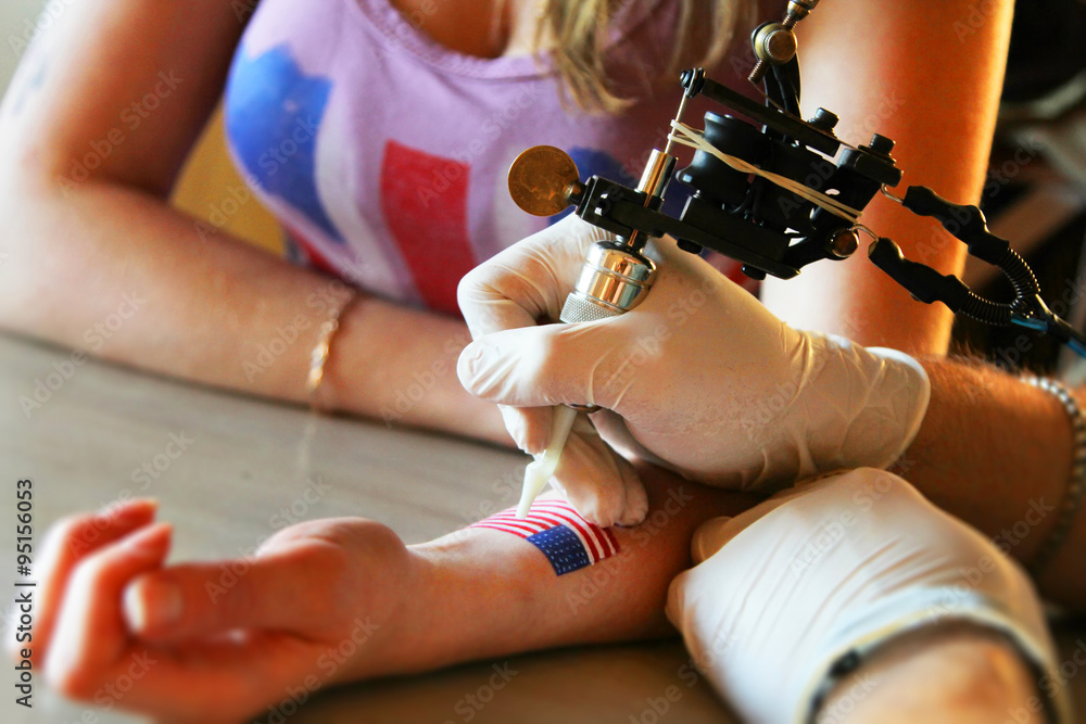 tattooer showing process of making a tattoo on young beautiful hipster woman with blonde hair hand. Tattoo design in the form of american flag