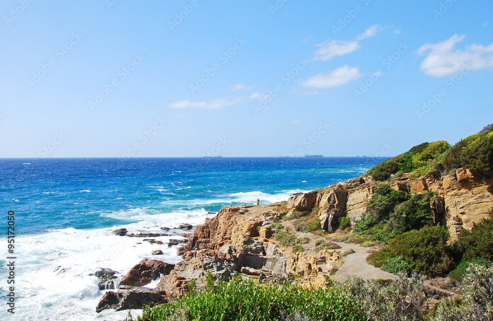 Rocky Italian coastline on a bright blue sky day, with blue water and some coastal vegetation. Strada del Vino Costa degli Etruschi.