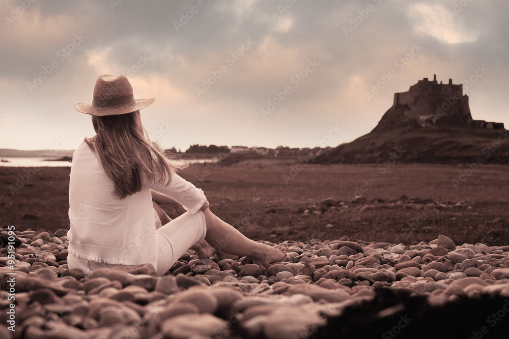 Woman sits on a pebble beach looking back towards Lindisfarne Castle (Holy Island) Northumberland, UK.