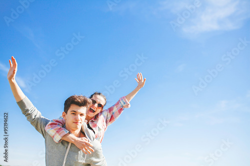 Teenager boy giving piggy back to his girlfriend. Piggyback couple on blue sky background.