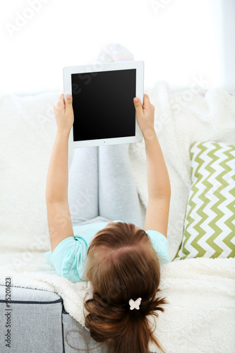 Beautiful little girl with digital tablet sitting on sofa, on home interior background