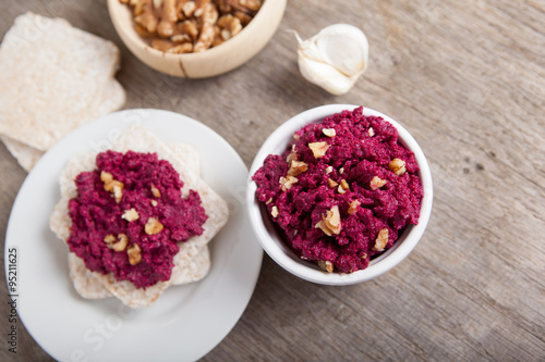 Beetroot pesto in a white bowl on a wooden table with garlic beetroot and rice cakes on the back  selective focus