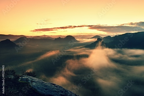 Warm misty autumn land in red colorful vapor. Rocky gulch full of heavy fog. Sun is hidden in colorful mist.