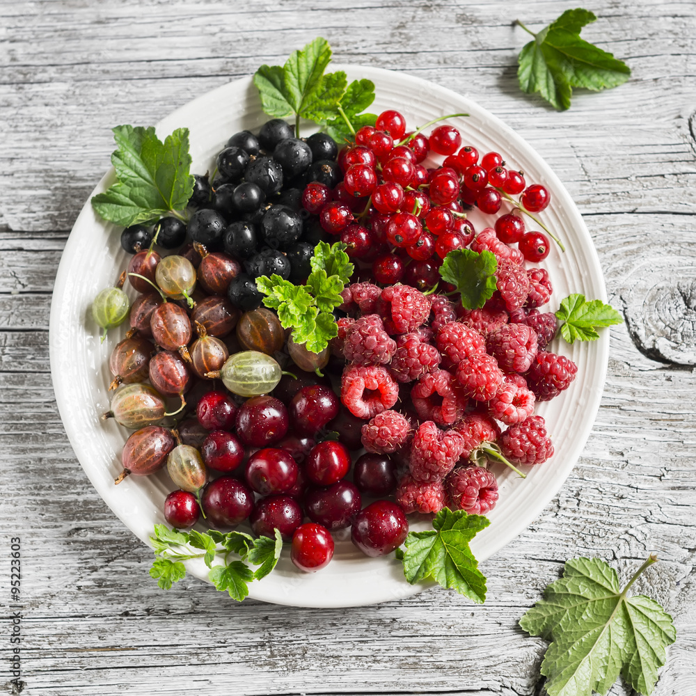 berries - raspberries, gooseberries, red currants, cherries, black currants on a white plate on a light wooden background berries - raspberries, gooseberries, red currants, cherries, black currants on a white plate on a light wooden background