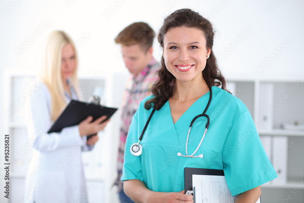 Beautiful young smiling female doctor standing at hospital with doctor and patient on the background. Medical concept. Beautiful young smiling female doctor standing at hospital with doctor and patient on the background. Medical concept.