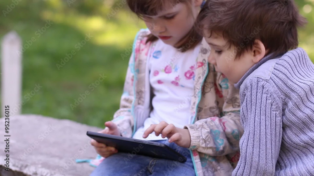 Little girl plays with tablet PC and brother looks at it in summer park
Little girl plays with tablet PC and brother looks at it in summer park