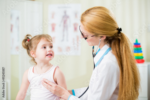 Pediatrician woman examining cute little girl with stethoscope. Kid happily smiling and looking at the doctor