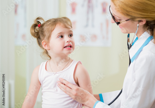 Pediatrician woman examining cute little girl with stethoscope. Kid happily smiling and looking at the doctor