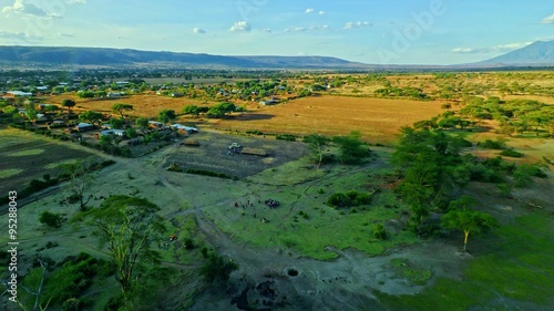 Aerial of African people in a remote bush village in Rift Valley of Tanzania, gathered by a water well well at sunset with vast vista view of the village houses, hills and fields in the distance
