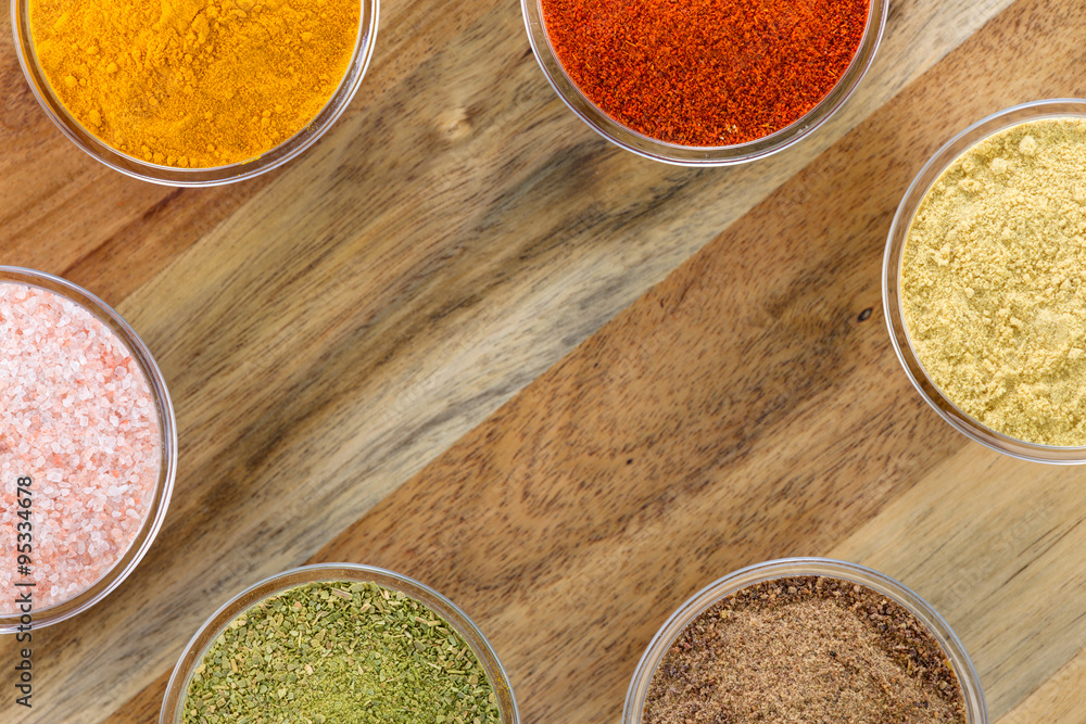 The powders of: bay leaf, turmeric, mustard, red paprika and nutmeg are poured in the glass bowls on the wooden background. The powders of: bay leaf, turmeric, mustard, red paprika and nutmeg are poured in the glass bowls on the wooden background.