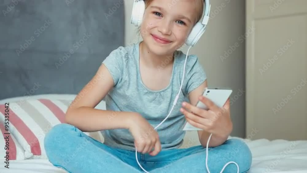 Child girl sitting resting on the bed and listening music in headphones from smartphone cell phone and dancing Child girl sitting resting on the bed and listening music in headphones from smartphone cell phone and dancing