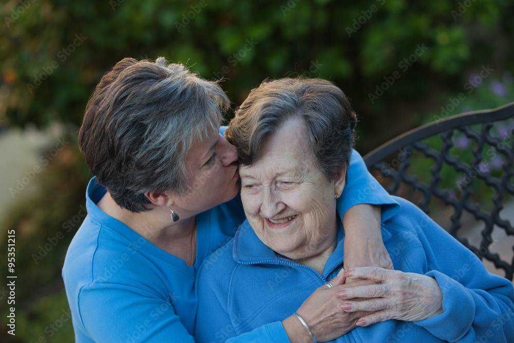 Daughter embraces mother and kisses her on the forehead. They are in a garden setting, are holding hands and wearing the same color blue shirts. A tender moment between a mother and daughter. Daughter embraces mother and kisses her on the forehead. They are in a garden setting, are holding hands and wearing the same color blue shirts. A tender moment between a mother and daughter.