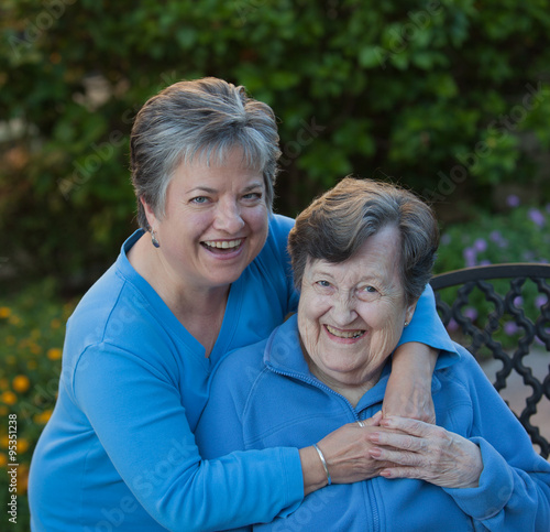 Daughter and mother are laughing in the garden. They are embracing and holding hands, sitting on a wrought iron bench, wearing the same color blue shirts. © jdpphoto15
