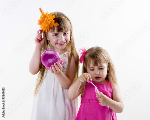  The older sister on the white dress takes care of the younger on the pink dress combing her hair and gets lipstick on her lips isolated on the white background