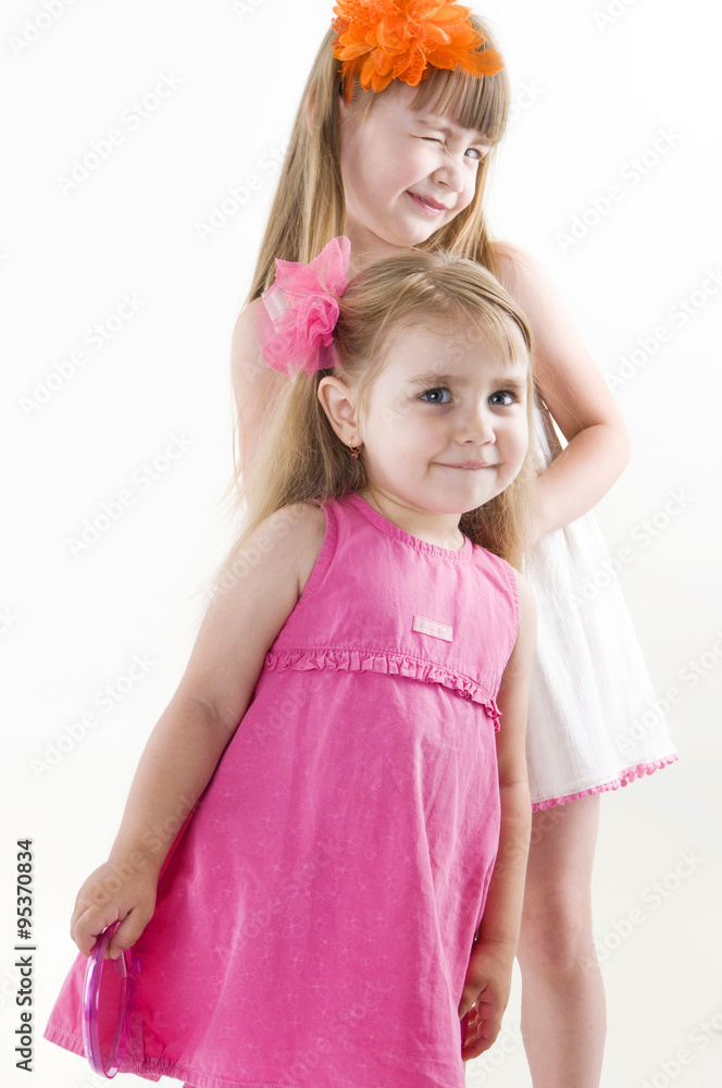 The older sister on the white dress takes care of the younger on the pink dress combing her hair and gets lipstick on her lips isolated on the white background The older sister on the white dress takes care of the younger on the pink dress combing her hair and gets lipstick on her lips isolated on the white background