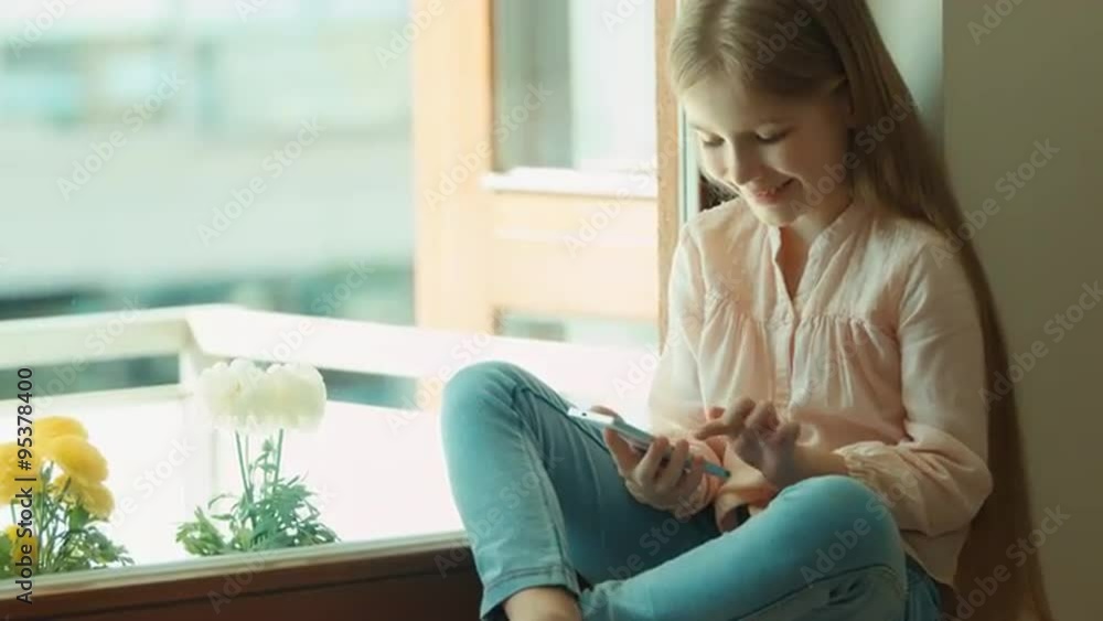 Child using mobile phone and smiling. Looking at camera. Shows ok. Thumbs up. Girl sitting on the window sill Child using mobile phone and smiling. Looking at camera. Shows ok. Thumbs up. Girl sitting on the window sill