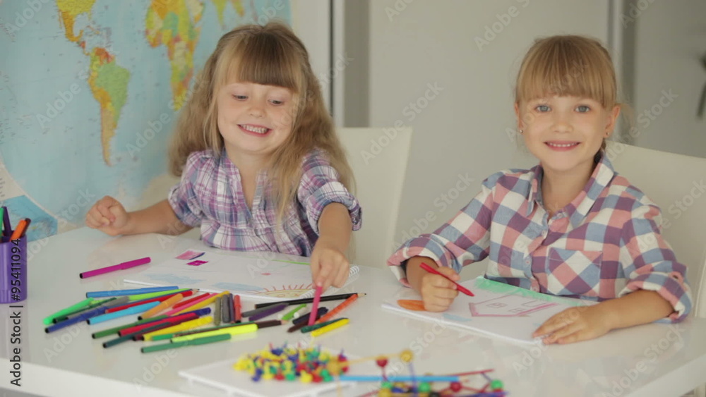 Two cute little girls sitting at table drawing with colored pencils and smiling Two cute little girls sitting at table drawing with colored pencils and smiling
