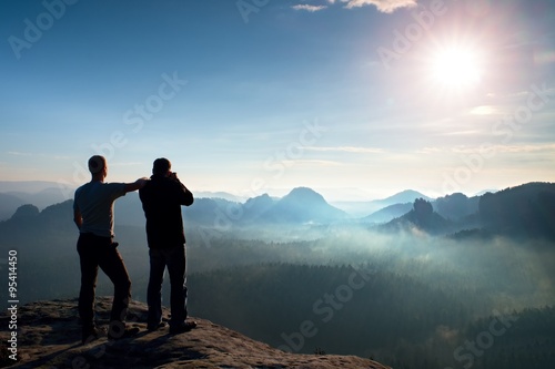 Two friends. Hiker thinking and photo enthusiast takes photos stay on cliff. Dreamy fogy landscape, blue misty sunrise in a beautiful valley below