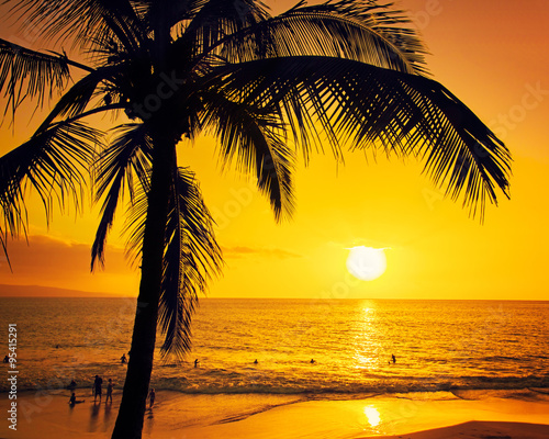 Tropical island sunset with silhouette of palm trees, hot summer day vacation background, golden sky with sun setting over horizon, people swimming in the ocean at sunset