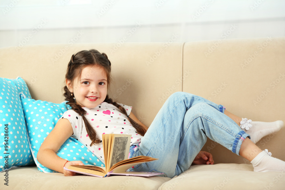 Beautiful little girl sitting on sofa with book, on home interior background Beautiful little girl sitting on sofa with book, on home interior background