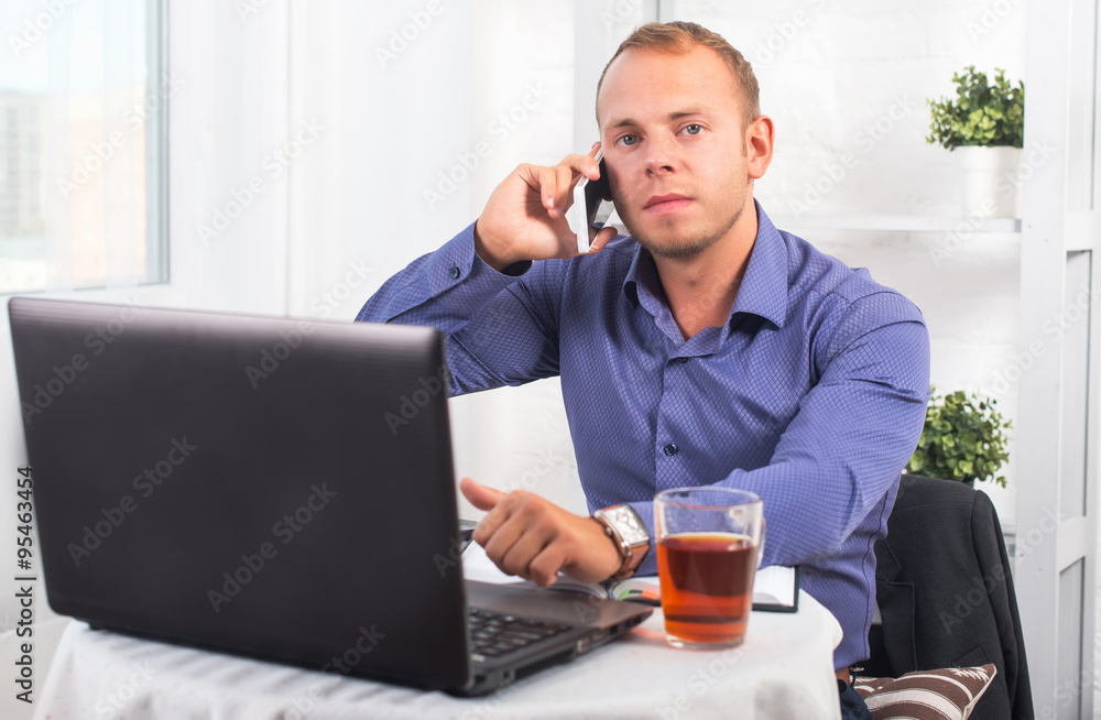 Young businessman working in office, sitting at a table, looking straight and talking on the phone Young businessman working in office, sitting at a table, looking straight and talking on the phone
