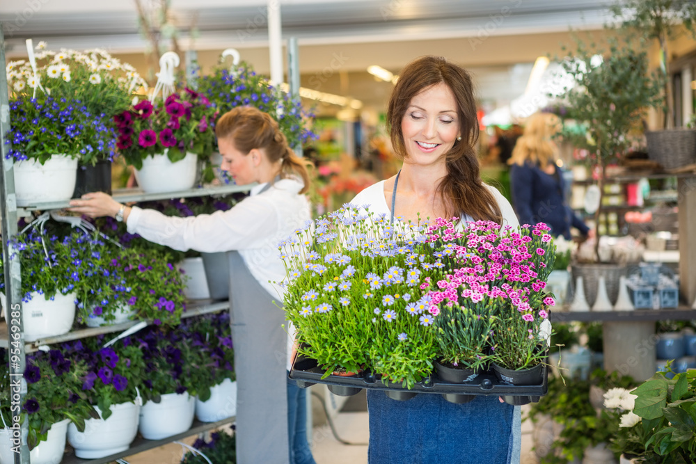 Smiling Female Worker Carrying Crate Of Flower Plants In Shop