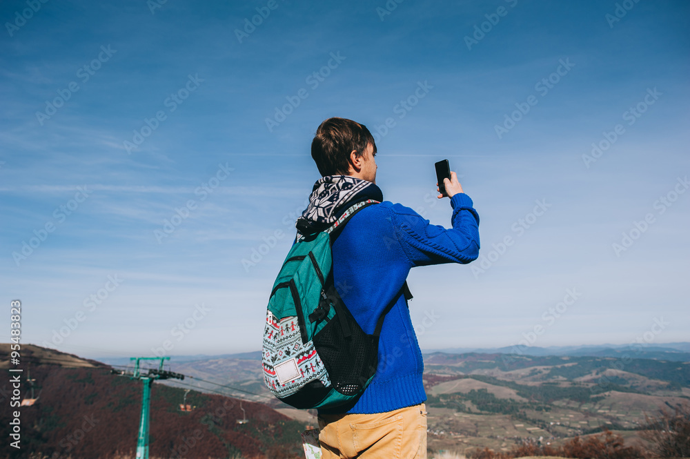 Young hipster taking photo by smart-phone on the peak of mountai